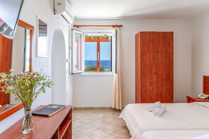 A hotel room with a bed, a desk with a laptop, a wooden wardrobe, a window with a view of the sea, and a vase with flowers. - Holiday Home 'Anemologio 202 Studio Tinos' with Sea View, Wi-Fi and Air Conditioning (Laouti)