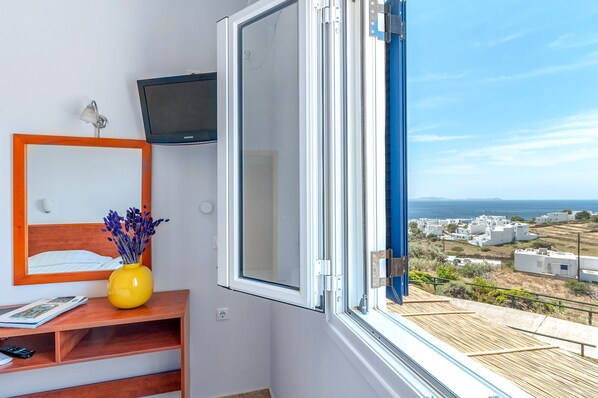 Une chambre avec un lit, une télévision, une fenêtre donnant sur des immeubles et un vase rempli de fleurs.
