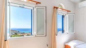 A room with a bed, a table with a bottle and glasses, and a view of the sea and buildings.