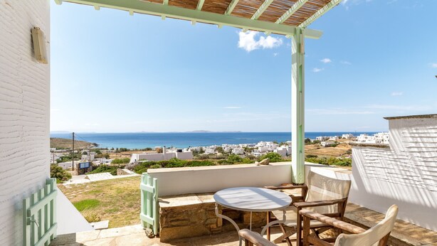 A terrace with a table and chairs, overlooking a coastal town with white buildings and a clear sky. - Holiday Home 'Anemologio 102 Tinos' with Sea View, Wi-Fi and Air Conditioning (Laouti)