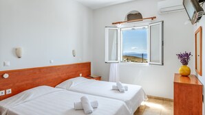 A hotel room with two beds, a wooden headboard, a window with an open view, a wooden nightstand, and a yellow vase with flowers. - Holiday Home 'Anemologio 102 Tinos' with Sea View, Wi-Fi and Air Conditioning (Laouti)