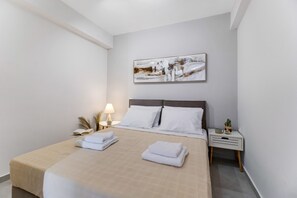 A neatly made bed with white linens, a bedside lamp, a small table with a plant, and a painting on the wall.