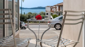 A balcony with two white chairs, a small table with a red flower, and a view of the sea and buildings. - Studio Apartment 'Mella Roa Teal' with Shared Terrace, Wi-Fi and Air Conditioning (Lefkada)