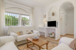 A living room with a sofa, a TV on a stand, a table with a bowl, and a window with curtains.