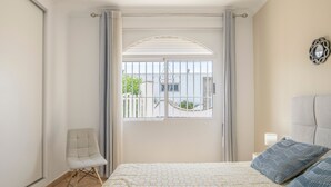 A bedroom with a bed, a chair, a window with curtains, and a nightstand with a lamp. - Holiday Home 'Chalet Fuente2 Shared Pool Family' with Shared Pool and Air Conditioning (Conil de la Frontera)