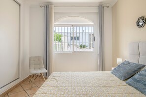 A bedroom with a bed, a chair, a window with curtains, and a nightstand with a lamp.