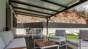 Un patio exterior con un sofá blanco, sillas grises, una mesa de madera y vistas a una ladera rocosa.