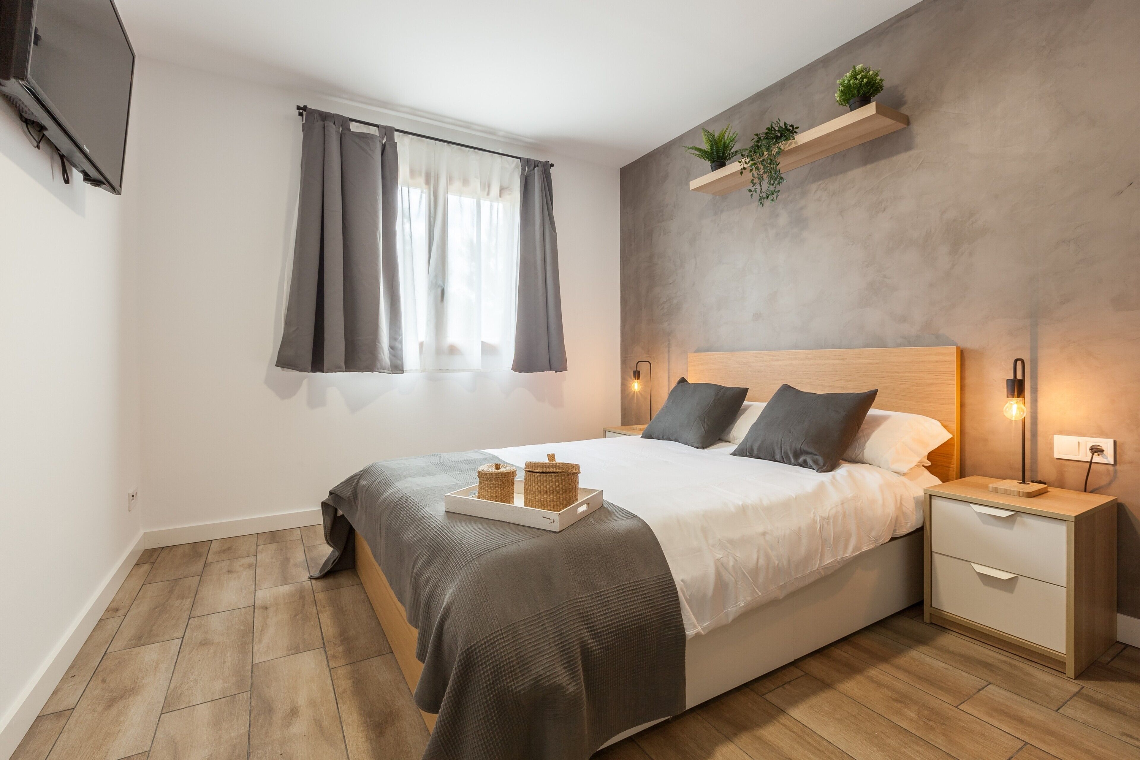 Une chambre à coucher comprenant un lit, des tables de chevet, une télévision, une fenêtre avec des rideaux et une étagère avec des plantes.