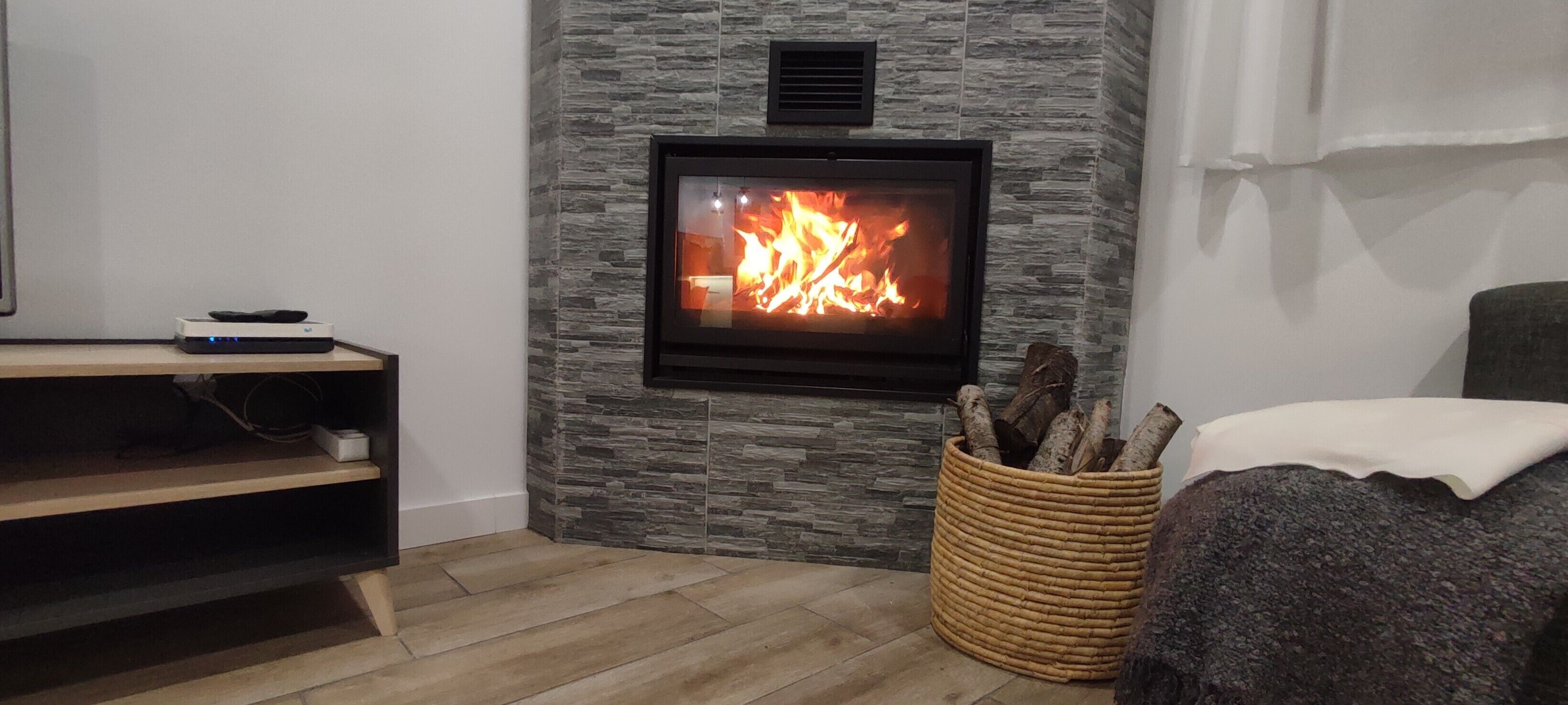 Un saló amb una paret de pedra, una llar de foc, una televisió, un paviment de fusta i una cistella de vimet plena de llenya.
