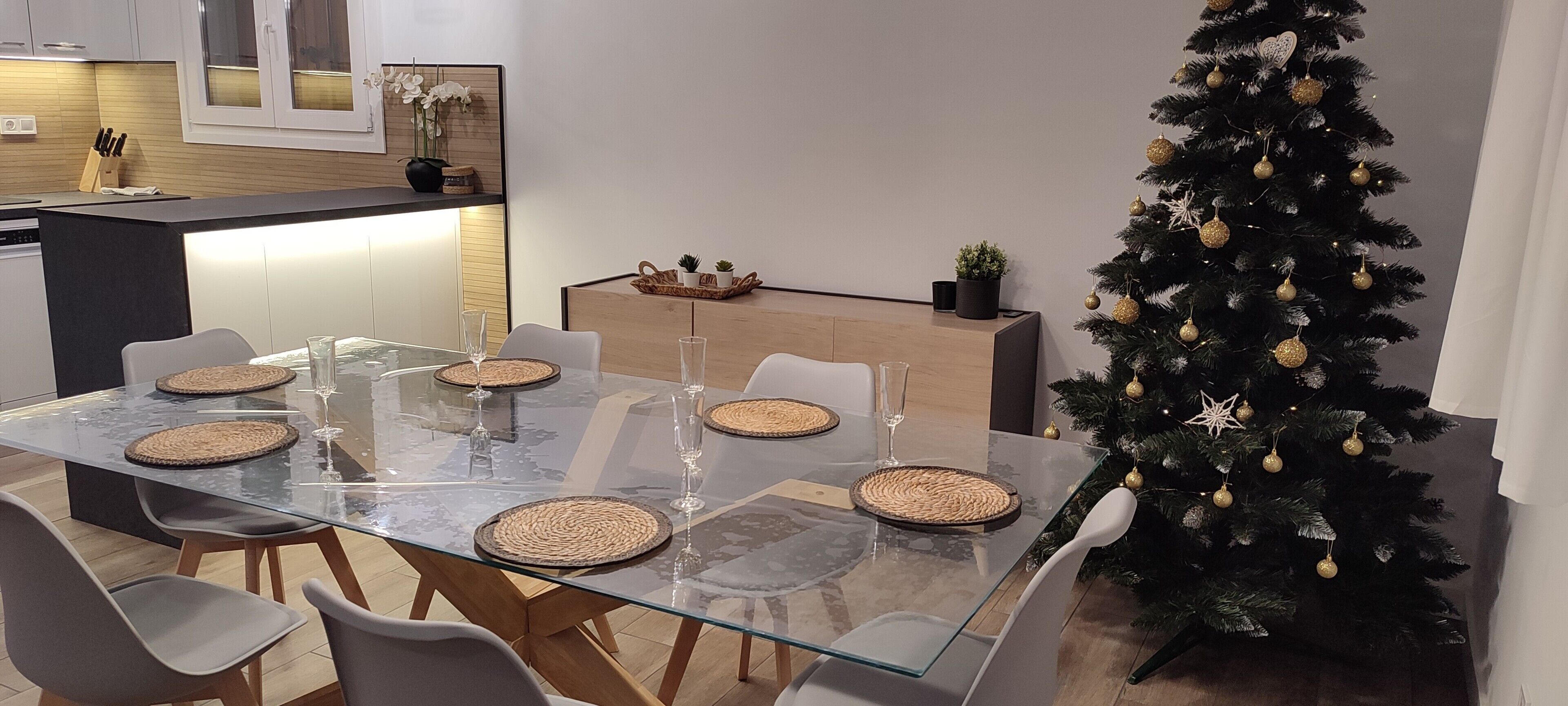 Un comedor moderno con una mesa de vidrio, sillas blancas y un árbol de Navidad.