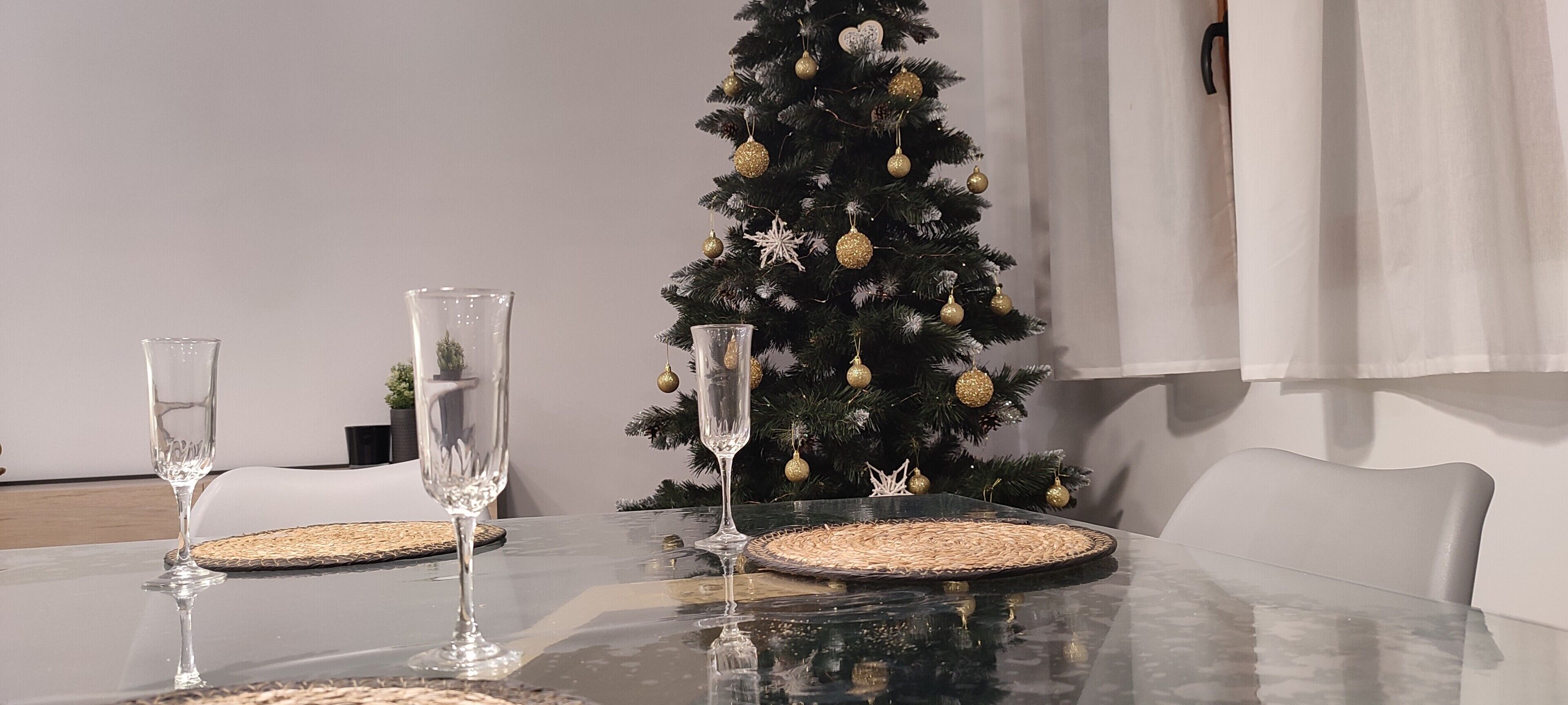 Una mesa de comedor con dos vasos transparentes, un posavasos tejido y un árbol de Navidad con adornos dorados.