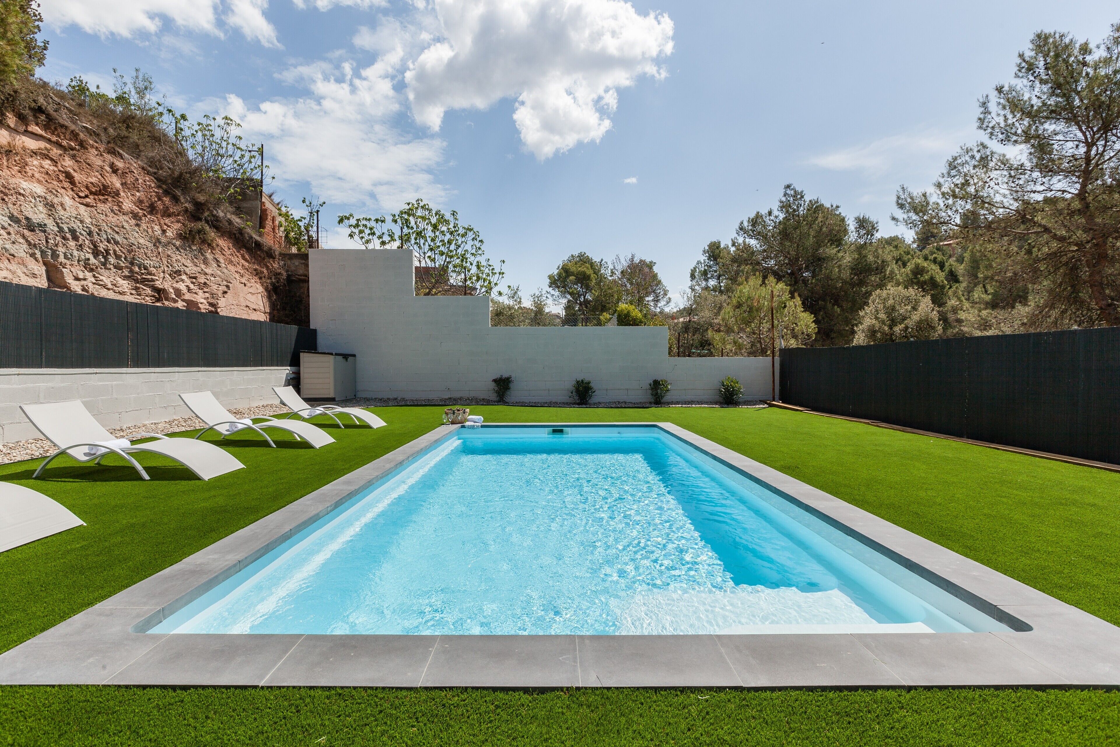 Una piscina rectangular con superficie de agua azul cristalina, rodeada por un césped cuidado y un bajo muro de piedra.