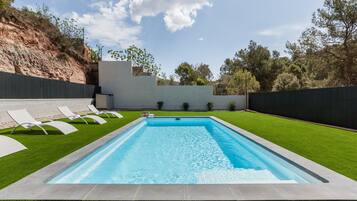 Una piscina rectangular con superficie de agua azul cristalina, rodeada por un césped cuidado y un bajo muro de piedra.