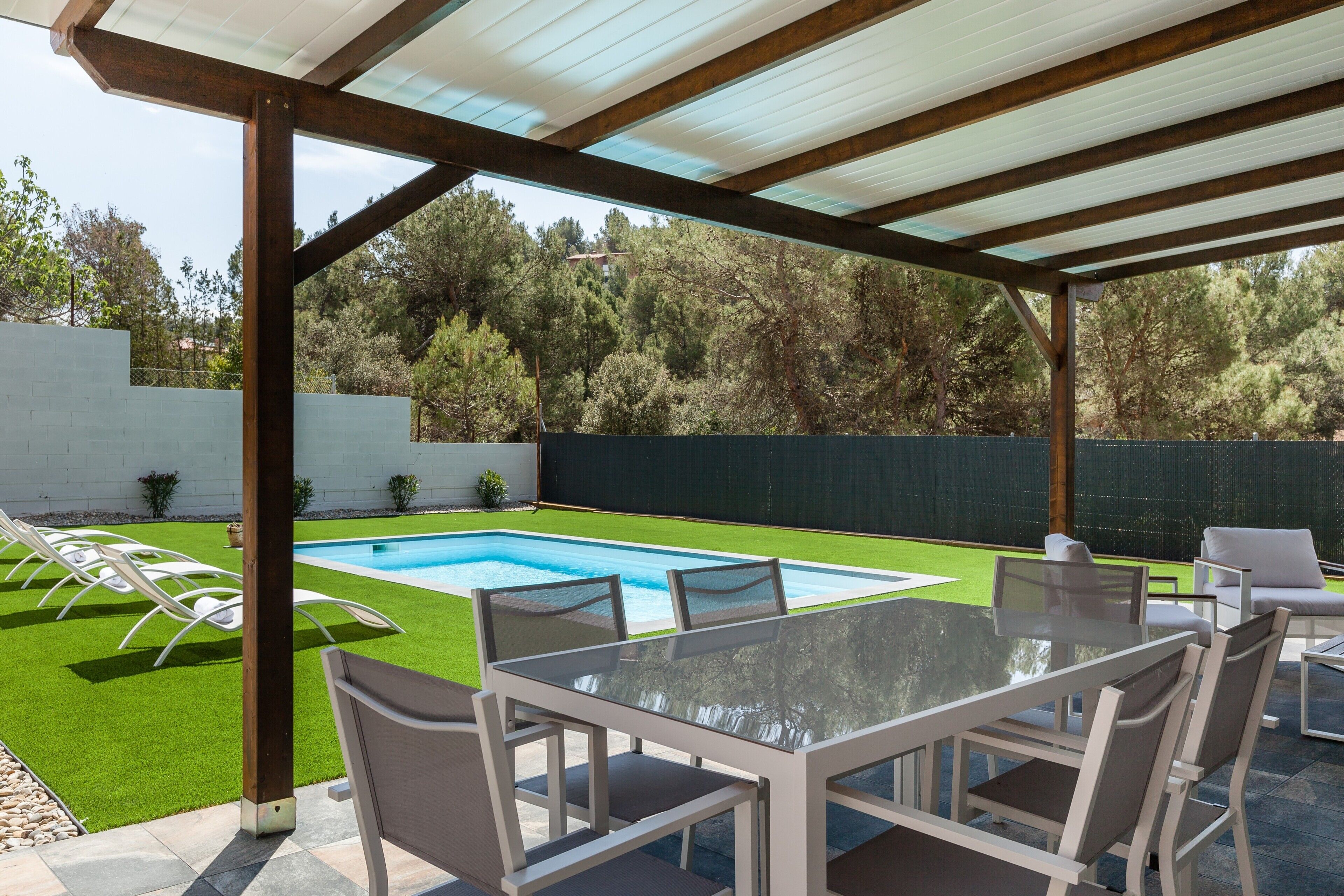 A covered outdoor area with a pool, a table with chairs, and a view of a green lawn and trees.