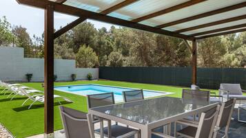 A covered outdoor area with a pool, a table with chairs, and a view of a green lawn and trees.