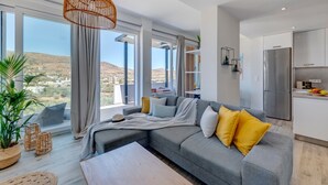 En moderne stue med en grå sofa, gule puder og udsigt til et bølgende landskab gennem store vinduer.