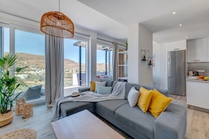 Un salon moderne avec un canapé gris, des coussins jaunes et une vue sur un paysage vallonné, visible à travers de grandes fenêtres.