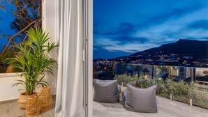 En balkon med udsigt over en by i skumringen, en potteplante og et hvidt gardin.