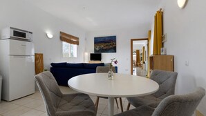 A compact living space with a kitchenette, a dining area with a round table and four chairs, a blue sofa, a flat-screen TV, and a painting on the wall.