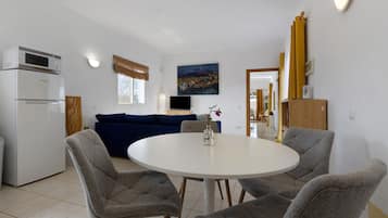 Ein kompakter Wohnbereich mit Küchenzeile, einer Essecke mit rundem Tisch und vier Stühlen, einem blauen Sofa, einem Flachbildfernseher und einem Bild an der Wand.
