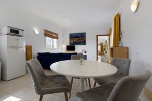 Un espace de vie compact comprenant un coin cuisine, un coin repas avec une table ronde et quatre chaises, un canapé bleu, un téléviseur à écran plat et un tableau accroché au mur.
