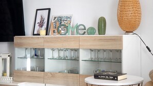Un salon moderne avec un meuble vitré où sont exposés des verres à vin, un cactus, des œuvres d’art encadrées et des lettres décoratives qui forment le mot « Accueil ».