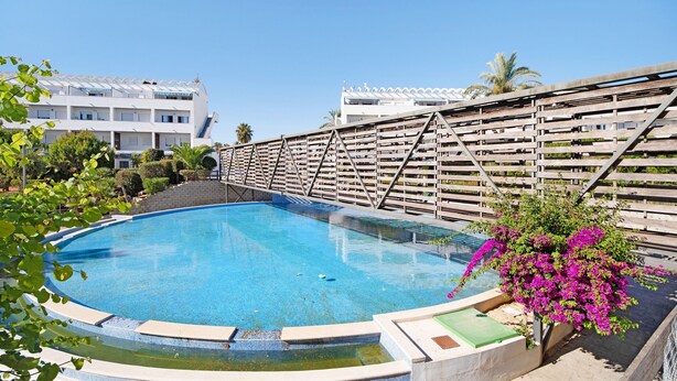 Une piscine dotée d’une terrasse en bois, entourée de verdure, avec un bâtiment visible au loin.