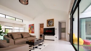 A modern living room with a sofa, a coffee table, and a flat-screen TV displaying 'NETFLIX'. There is a large window with curtains, a painting on the wall, and a glass door leading to an outdoor area with colorful cushions. - Pool villa at Kamala Regent by Lofty (Kamala)