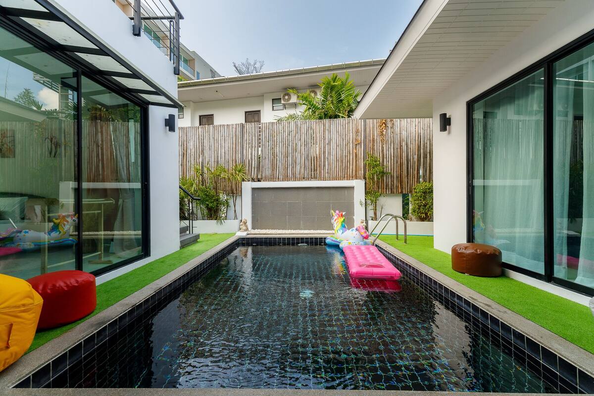 A modern outdoor swimming pool with a wooden fence, green artificial turf, and colorful cushions.