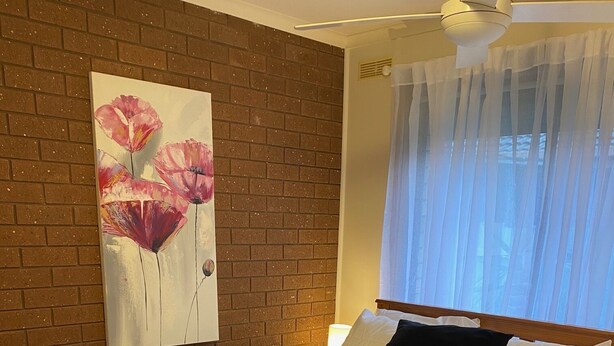 A bedroom with a brick wall, a painting of flowers, a bed with white and black pillows, and a window with sheer curtains. - Quiet and comfortable 2 bedroom apartment, 65 inch Smart TV (Wodonga)