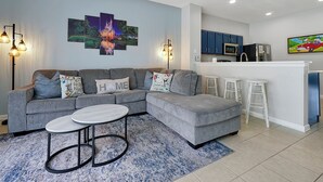 A modern living room with a grey sectional sofa, a round coffee table, and a kitchen area with blue cabinets and a white countertop. - Home minutes to Disney with private pool, arcade, waterslides, gated resort (Kissimmee)