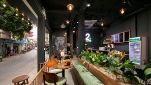 Un café intérieur avec des meubles en bois, un banc recouvert d’un tissu vert et une variété de plantes.