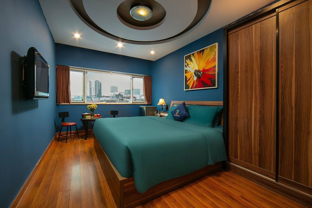 A hotel room with a bed, a desk with a chair, a window with curtains, a painting, and a TV on the wall.