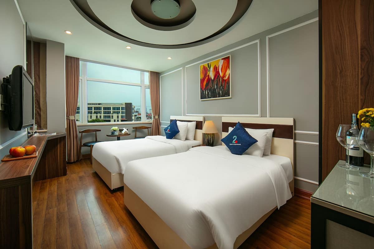 A hotel room with two beds, a desk, a TV, and a window with a city view.