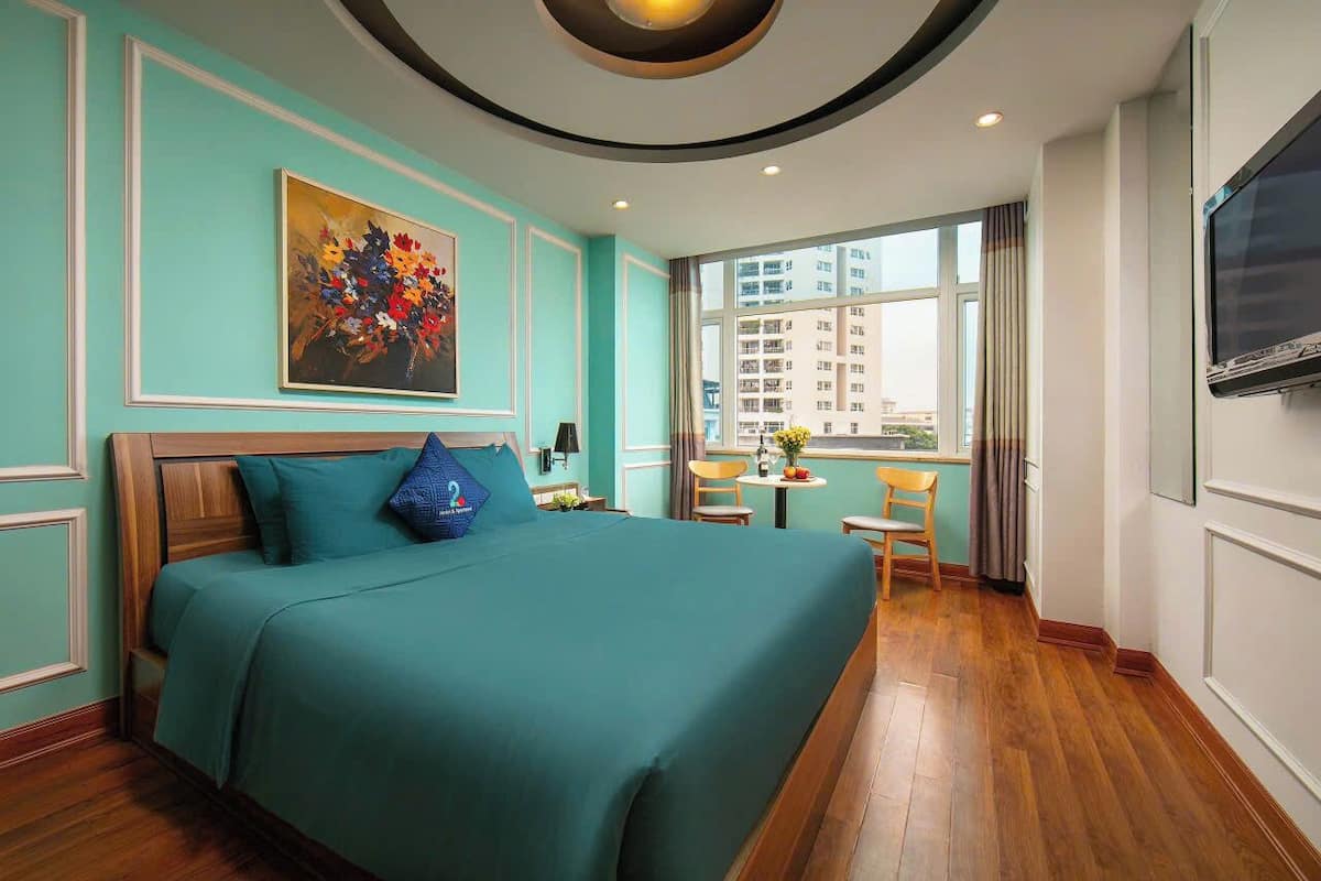 A hotel room with a large bed, a painting on the wall, a window with a view of buildings, and a TV mounted on the wall.