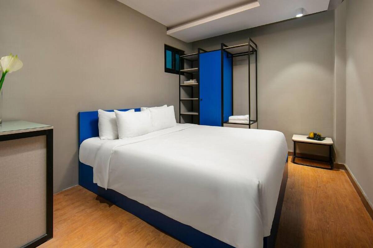 A modern hotel room with a neatly made bed, a small bedside table, a desk with a vase of flowers, and a blue wardrobe.