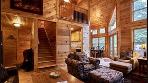 A log cabin interior with a staircase, two sofas, a dining area, and large windows. - Mountain Memories - Cabin near Blue Ridge Parkway (Boone)