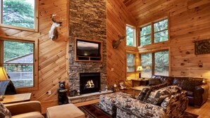 A log cabin interior with a stone fireplace, wooden walls, a television, and a patterned sofa. - Mountain Memories - Cabin near Blue Ridge Parkway (Boone)