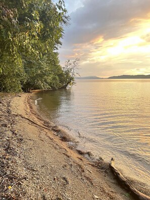 Une plage au rivage de galets, bordée d’une rangée d’arbres, et une mer calme sous un ciel au coucher du soleil.