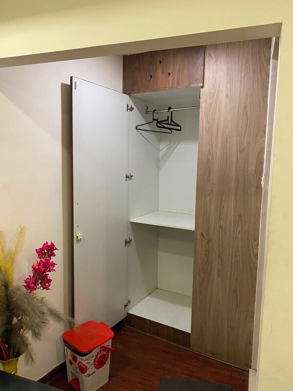 Quarto com guarda-roupa de madeira, porta branca e uma pequena lixeira com tampa vermelha.
