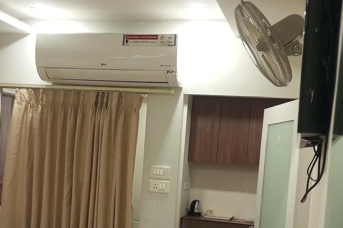 A hotel room with a bed, an air conditioner, a fan, a small table with a kettle, and a cabinet.