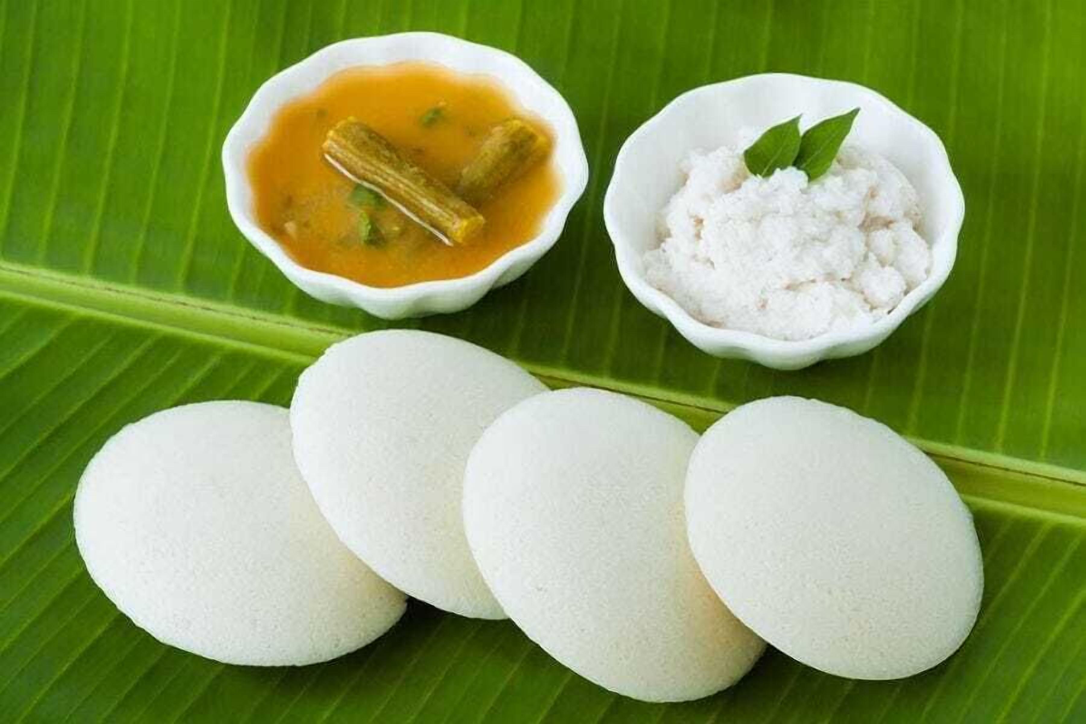 Ein Teller Idlis mit einer Schüssel Kokosnuss-Chutney und Sambar, sowie eine Schüssel Kokosraspeln auf einem Bananenblatt.