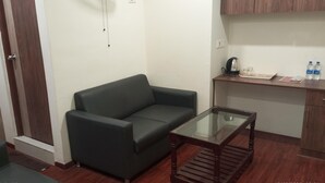 Una sala de estar con un sofá verde oscuro, una mesa de centro de vidrio, un mueble de madera y una pequeña área de cocina con una tetera y botellas de agua.