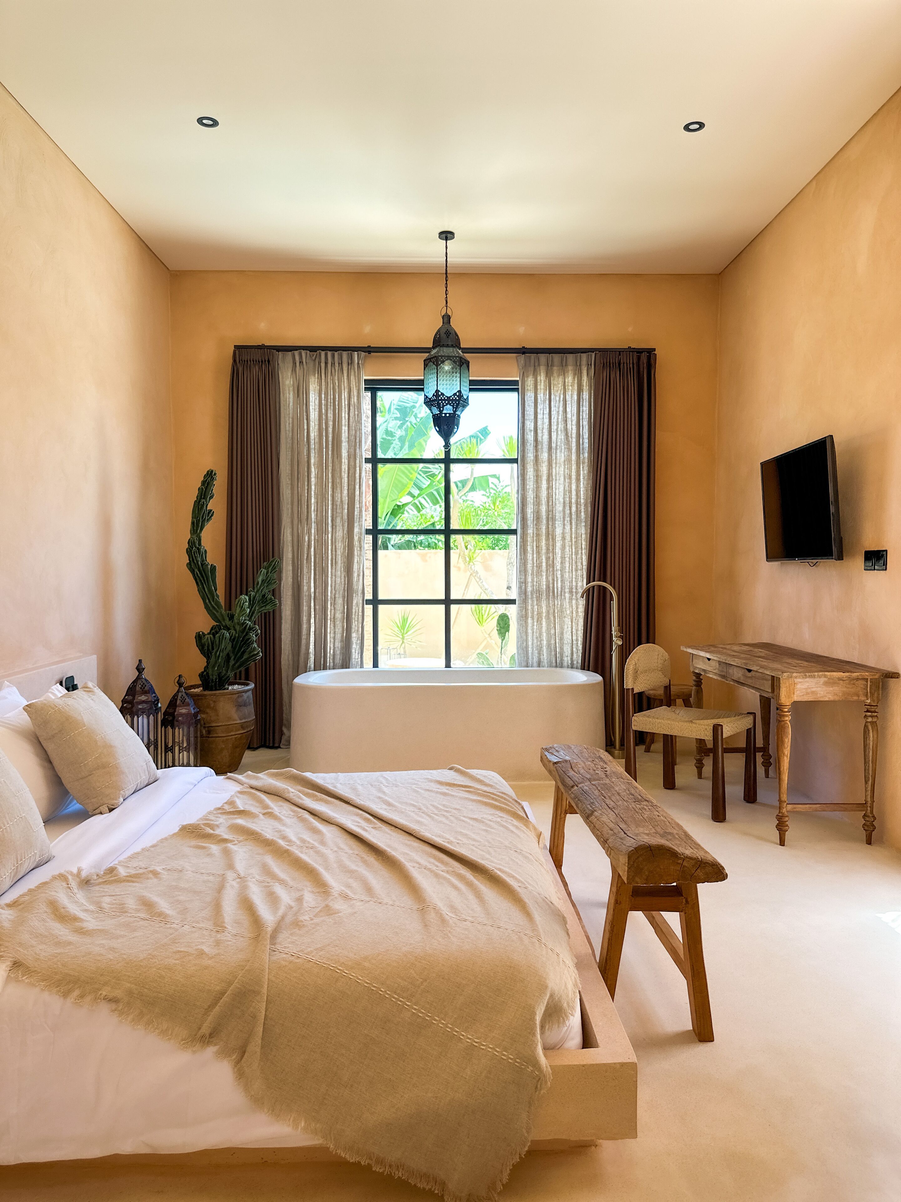 A bedroom with a large bed, a wooden bench, a desk, a chair, a television, and a window with curtains.