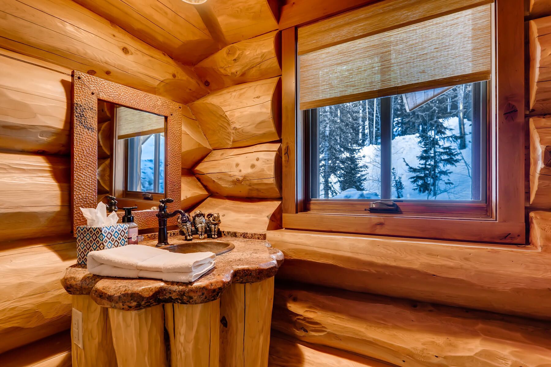 Een houten badkamer in een blokhut, met een houten wastafel, een spiegel en een raam met uitzicht op besneeuwde bomen.
