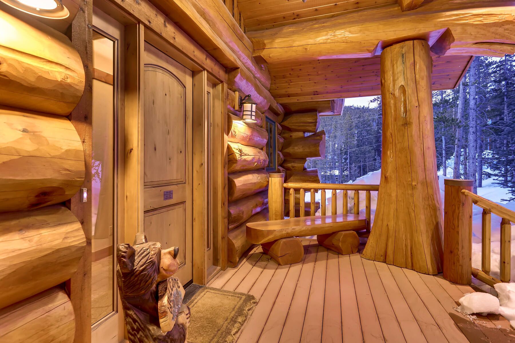A log cabin with a wooden deck, a bench, and a view of the snowy forest.