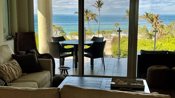 Ein Zimmer mit einer Couch, einem Tisch und Stühlen, vor einem großen Fenster mit Blick auf einen Strand mit Palmen und dem Meer.