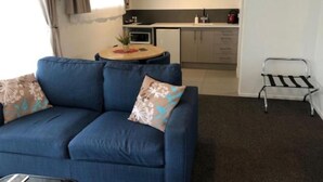 En kompakt opholdsstue med en sofa, et lille rundt bord, et tekøkken og en metalstol.