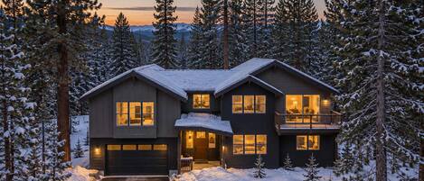 A two-story house with a garage, surrounded by snow-covered trees.