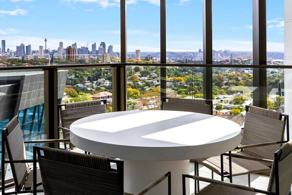 A balcony with a round table and chairs, overlooking a cityscape with buildings and a bridge.